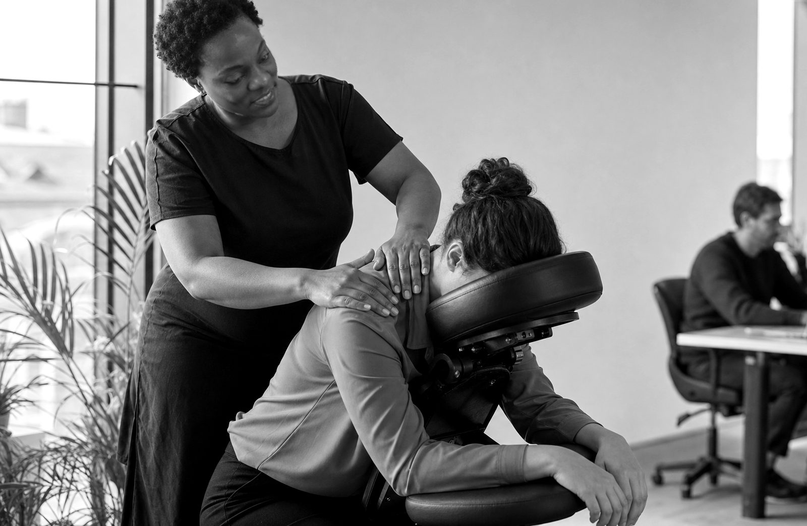 Séance de massage assis sur chaise ergonomique dans un bureau, une salariée habillée reçoit un massage des épaules pendant que ses collègues travaillent en arrière-plan.