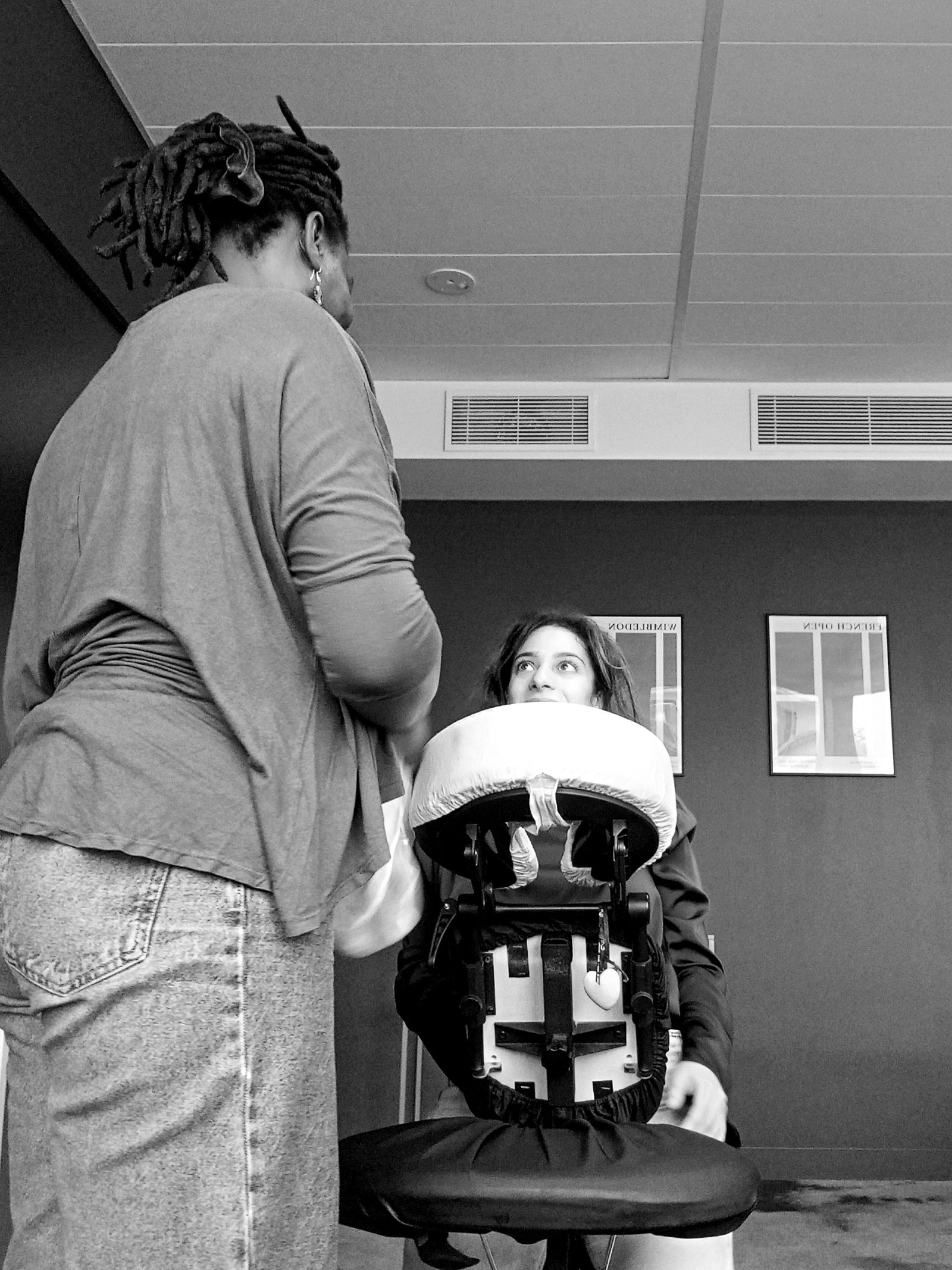 La praticienne masse les épaules d’une salariée installée sur la chaise ergonomique, qui lève les yeux vers elle en souriant.